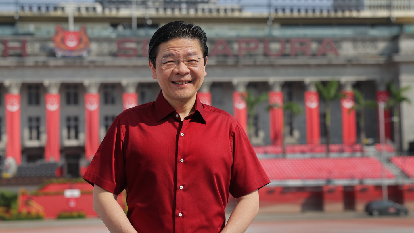 Man in deep red shirt at parade venue; civic building and red banners in background.